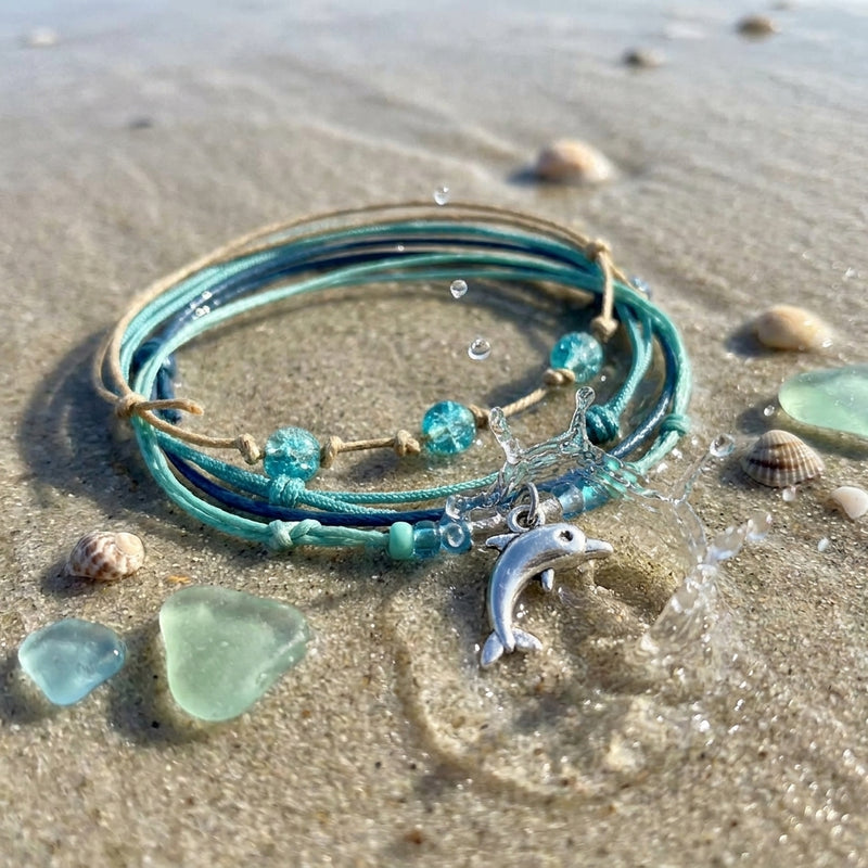 Green Dolphin Bracelet Style Pack on wet sand with sea glass and shells, the dolphin charm catching a splash