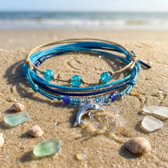 Blue Dolphin Bracelet Style Pack on beach sand with sea glass and shells, the dolphin charm by a small splash and the surf behind