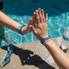 Two friends high-fiving by a pool while wearing matching blue 67 string bracelets from O Yeah Gifts, lifestyle jewelry image of a waterproof 6-7 meme bracelet perfect for summer, 67 trend bracelet and school-safe friendship bracelet that makes kids ask what is 6 7 and what does 67 mean when they see the white 6 and 7 beads.