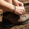 Close-up of a hiker tying brown leather boots in the woods while wearing a tan and brown 67 bracelet from O Yeah Gifts with white beads reading 6 and 7, outdoorsy 6-7 meme bracelet and gen alpha funny slang bracelet, six seven meme accessory that answers what does 6-7 mean for fans of the 6-7 TikTok trend, rugged yet school safe bracelet for teens, 67 trend jewelry that works as a six seven funny gift and 6-7 meme merchandise you can buy 6-7 bracelet online.