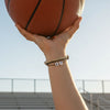Arm raised high holding a basketball in the air with stadium bleachers behind, wrist stacked with tan and gold 67 bracelets from O Yeah Gifts, spotlight on 6-7 meme bracelet as a winning IYKYK accessory, six seven meme jewelry for athletes who follow the 67 trend, gen alpha meme jewelry and viral gen alpha accessory that stays school safe, 6-7 meme merchandise and six seven funny gift you can buy 6-7 bracelet online, perfect for 6-7 TikTok trend fans.