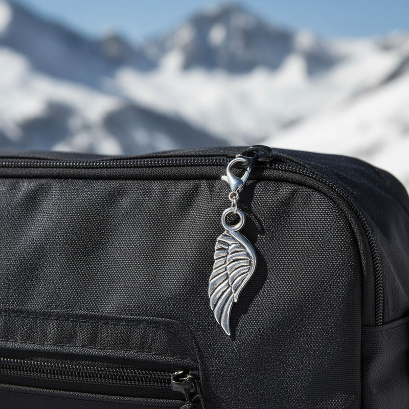 Silver angel wing clip charm clipped to the zipper of a black travel backpack with snowy mountains in the background, guardian angel zipper pull for ski trips and Arctic travel, winter travel bag accessory for suitcases, ski boot bags, kids backpacks and carry-ons, thoughtful holiday gift for travelers and families who love cold-weather adventures.