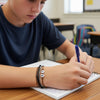 Teen boy in a classroom writing in a notebook while wearing black gray and brown 67 string bracelets with white number beads reading 6 and 7, school safe 6-7 meme bracelet and subtle six seven slang accessory, perfect gen alpha 6-7 jewelry for the 6-7 kid TikTok trend and Mr 6-7 Taylen Kinney fans, what does 6-7 mean meme answer on his wrist, viral 67 trend bracelet for everyday wear, stackable teen bracelets and school friendly accessory for bestie bracelets and tradeable bracelets, six seven funny gift.