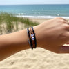 Outstretched arm with black and gray 67 bracelet stack from O Yeah Gifts in front of ocean waves and beach grass, simple 6-7 meme bracelet shown in a coastal lifestyle jewelry image, beachy fun fashion jewelry that stays on in the water, six seven meme jewelry for gen alpha kids who made the 6-7 TikTok trend huge, perfect answer when people online search “what is 6 7?” and “what does 67 mean?” and land on this 67 trend bracelet, school friendly accessory and 6-7 friendship bracelet gift.