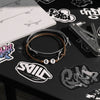 Flat lay jewelry image of a brown and gray 67 bracelet from O Yeah Gifts on a sticker-covered desk with a skate tool and doodled skateboard notebook, product photography jewelry shot of a 6-7 meme bracelet styled for skaters, perfect for online shoppers typing what is 6 7, what does 67 mean, 6-7 meme bracelet and buy 6-7 bracelet online.