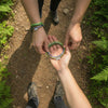 Top-down view of two hikers on a dirt trail holding a circular bundle of green 67 bracelets between them, flat lay meme jewelry style image and ai optimized product image for 6-7 meme bracelet packs from O Yeah Gifts; fun fashion jewelry and product photography jewelry for online shoppers literally searching “what is 6 7?” and “what does 67 mean?” as they discover six seven meme jewelry, gen alpha 6-7 bracelet bundles for tradeable bracelets, stackable teen bracelets and 6-7 meme merchandise, trend jewelry 