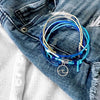 Stackable wave bracelet set with sea glass beads and silver wave charm, featuring adjustable string cord bracelets in ocean blue tones styled on denim and white top.