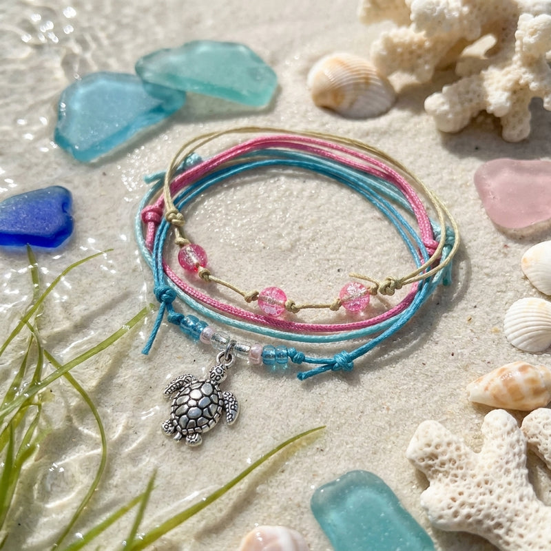 Sea Turtle Bracelet Style Pack Pink on wet sand with sea glass, shells and coral, the silver-tone turtle charm in front.