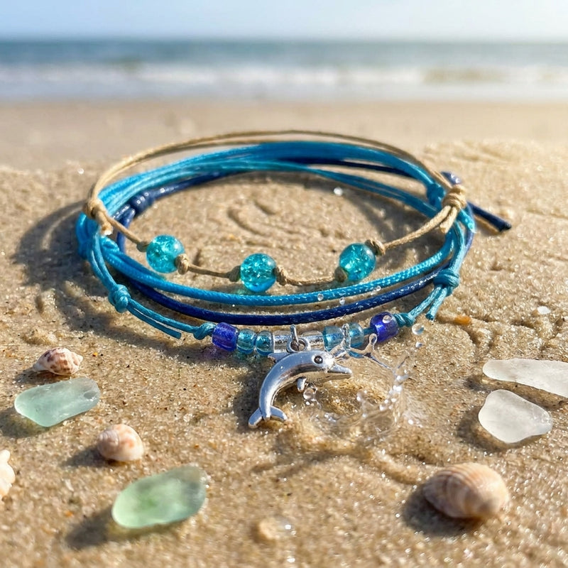 Blue Dolphin Bracelet Style Pack on beach sand with sea glass and shells, the dolphin charm by a small splash and the surf behind
