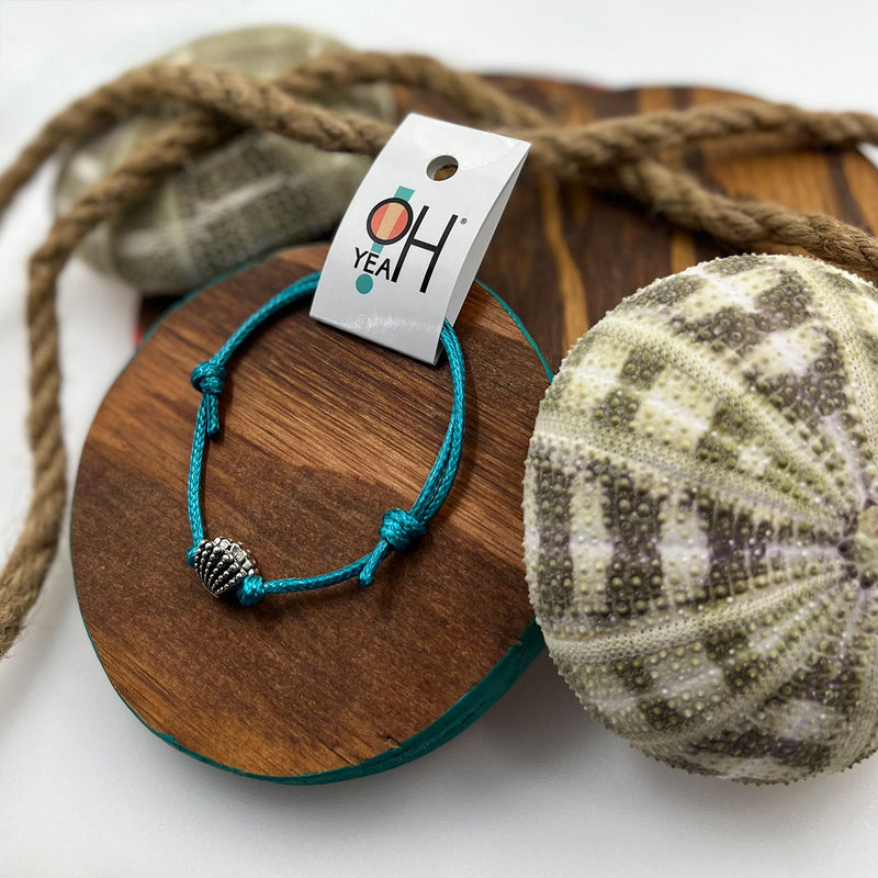 Silver shell charm bracelet on blue cord displayed on a wood board with sea-inspired decor from O Yeah Gifts.